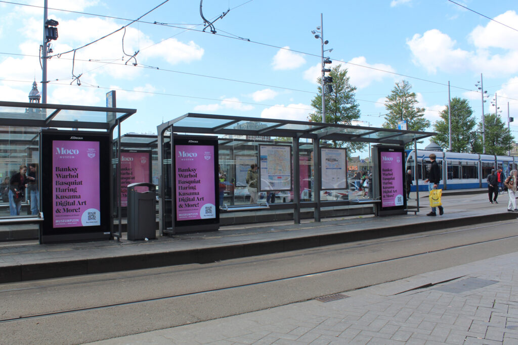 A tram stop in front of Amsterdam Central Station. Digital City Light Posters are displayed in the shelters, featuring eye-catching pink advertisements for the Moco Museum in Amsterdam.