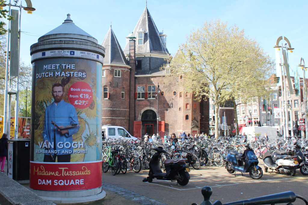 A large advertising column in Amsterdam. It is fully wrapped with a Madame Tussauds Amsterdam design. In the background, there are many bicycles, people and a large building.