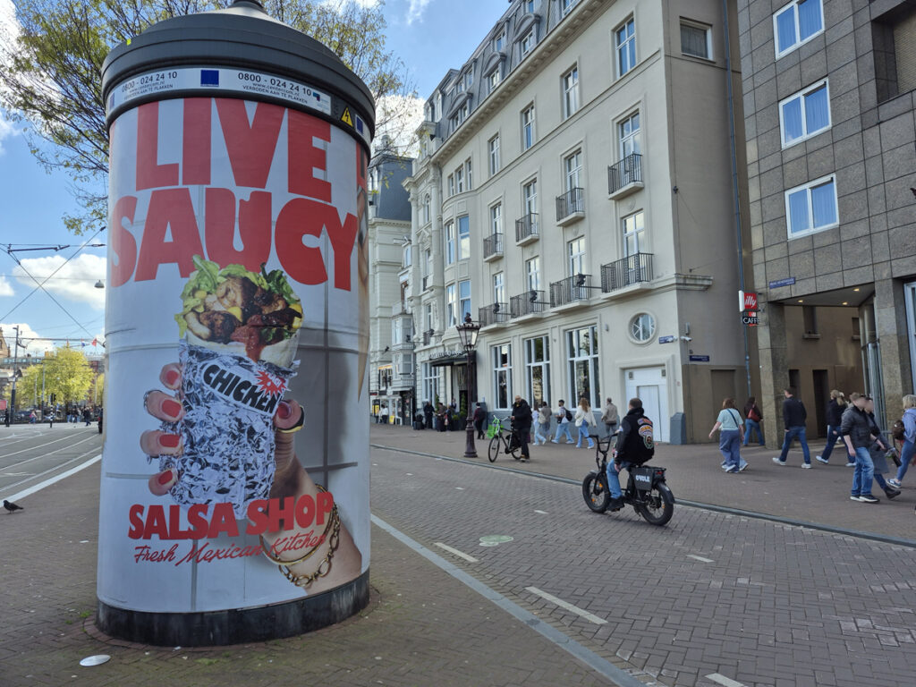 A large advertising column in the city of Amsterdam. The road is on the left, while the cycle path and pedestrian walkway are on the right. The column is fully wrapped with a design from Salsa Shop.