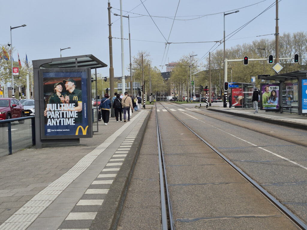 The tram stop at the RAI in Amsterdam. People are walking towards the exhibition centre. Analogue City Light Posters featuring advertising, including from McDonald’s, are displayed in the shelters.
