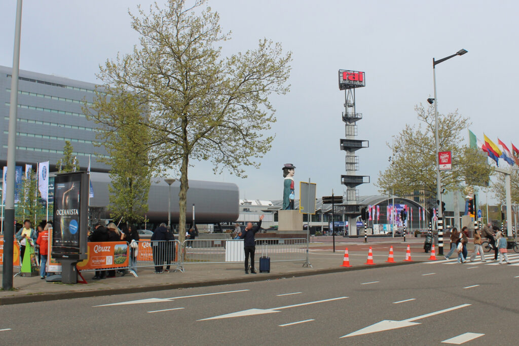 The RAI exhibition centre in Amsterdam. In the foreground, a digital City Light Poster is positioned directly along the road. People are crossing the street and waiting for a bus near the advertising display.