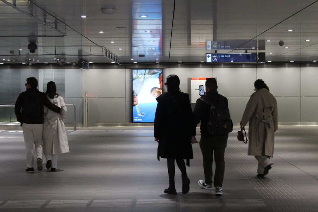 People are walking through Europaplein metro station directly towards a digital City Light Poster mounted on the wall. The LED illumination makes the outdoor advertising stand out.