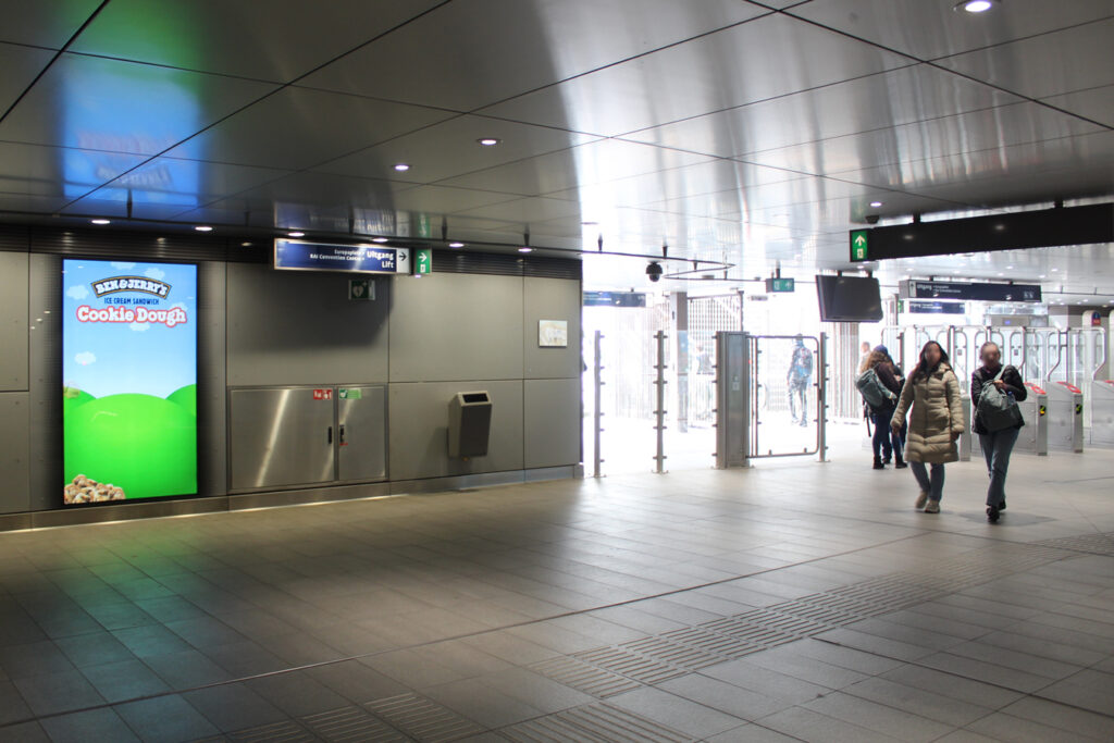 The concourse of the Europaplein subway station, with an exit leading to the RAI exhibition grounds. People are walking through the station. On the left wall hangs a digital City Light Poster displaying an outdoor advertisement for Ben & Jerry’s.