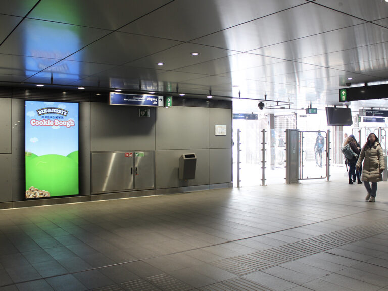 The concourse of the Europaplein subway station, with an exit leading to the RAI exhibition grounds. People are walking through the station. On the left wall hangs a digital City Light Poster displaying an outdoor advertisement for Ben & Jerry’s.