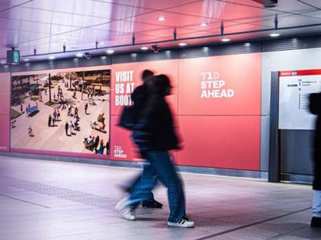 The large-format digital screen at Europaplein metro station near the RAI in Amsterdam. The wall surrounding the screen is wrapped to match the outdoor advertising displayed on the screen. The advertisement highlights a company’s trade show presence. People are walking by.