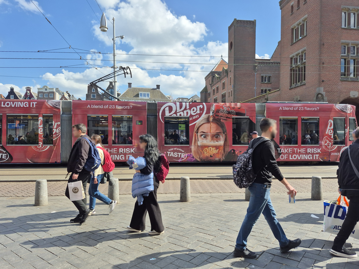 A tram travels through Amsterdam. People are walking in the foreground. The tram is fully wrapped in red Dr Pepper advertising, with only the windows left uncovered.