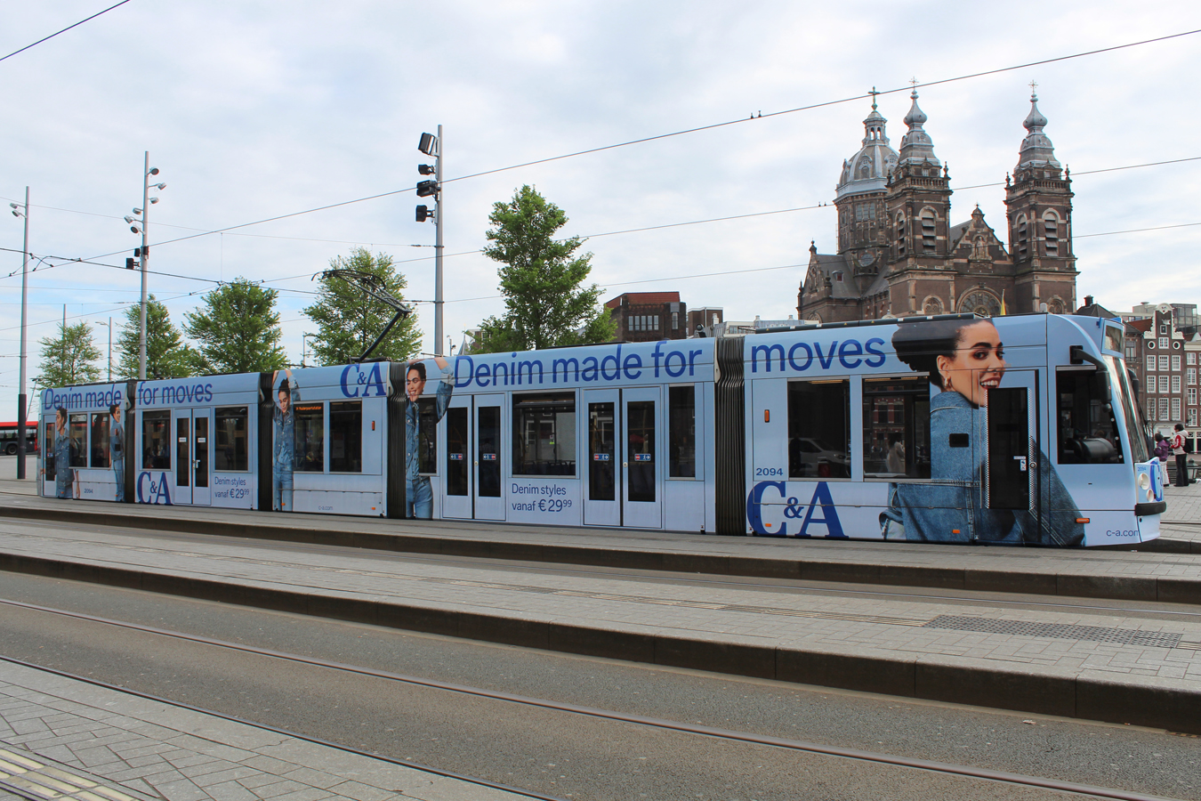 A tram fully wrapped in outdoor advertising stopped in front of Amsterdam Central Station. The advertising features a blue C&A design.