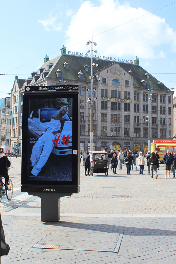 A digital City Light Poster in Amsterdam city centre. Many people are walking and cycling through the area. The screen displays outdoor advertising from H&M.