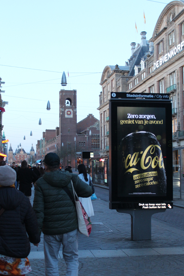 Digital City Light Poster on a street in Amsterdam. Many people are walking past. Coca Cola has booked outdoor advertising on the screen.
