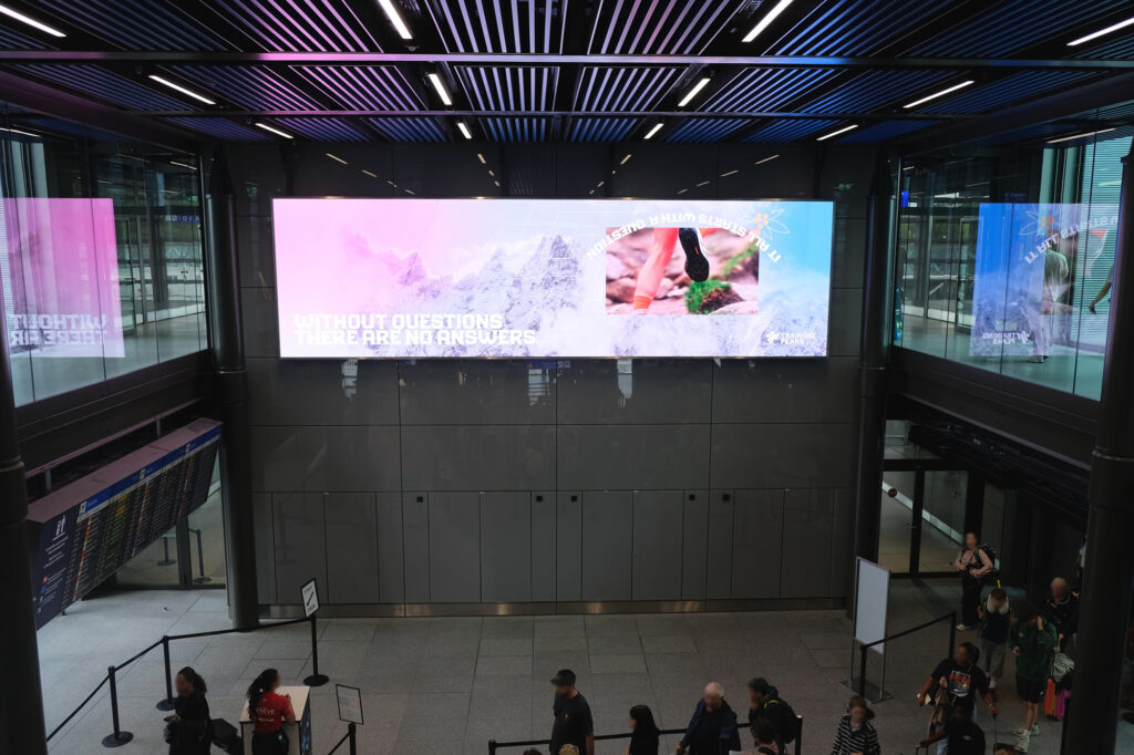 A large digital advertising display in an airport concourse. The display shows blue-and-pink TrainingPeaks ads featuring mountain scenes and a runner.
