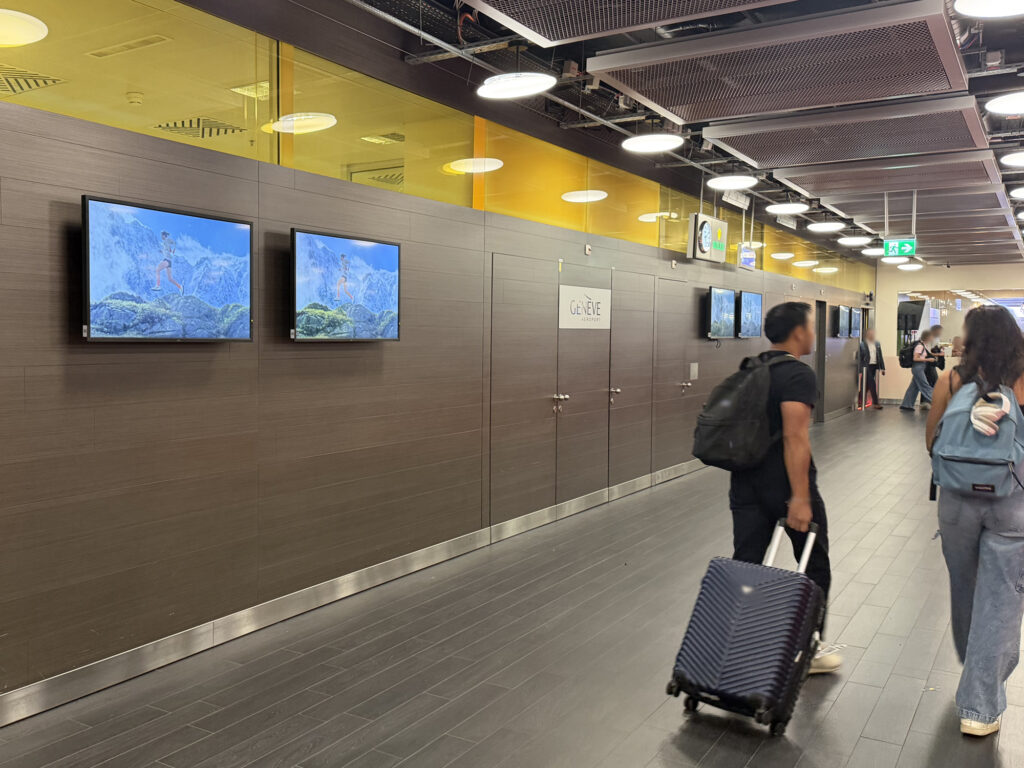 The arrivals hall at Geneva Airport. Six digital screens hang on the walls. They display synchronized advertisements. People walk by.