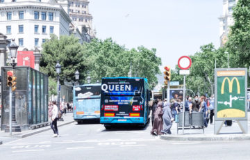 On the rear side of a bus at an intersection in Barcelona, you can see an advertisement for the rock band 'Queen.'
