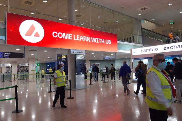 At Barcelona Airport, a large red ad shines on a screen hanging above the busy hall.