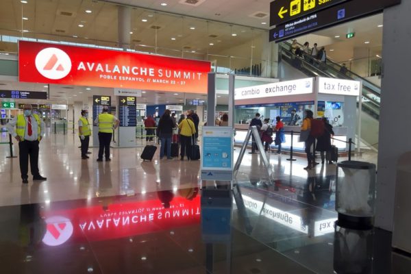 At Barcelona Airport, in a bustling hall, people wait as a large out-of-home screen displaying a striking red advertisement hangs prominently from the ceiling.