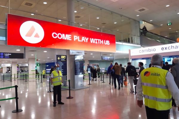 At Barcelona Airport, a busy hall filled with waiting travelers features a massive out-of-home screen showcasing a bold red advertisement, suspended from the ceiling.