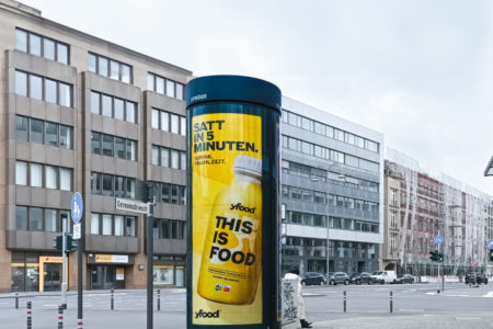 A City Light column in Cologne in front of a street intersection displays advertising from the company 'yFood'.