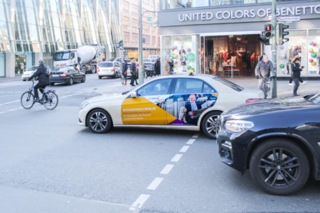 A taxi with a Door Cover advertising motif is at a street intersection in Cologne.