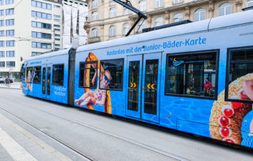 A tram is traveling through the city center of Frankfurt am Main with a full-cover motif promoting the public swimming pools in Frankfurt am Main.