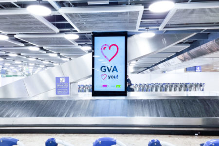 In front of the baggage claim at Geneva Airport, there is a digital City Light poster featuring a red heart. In the foreground, luggage carts are visible.