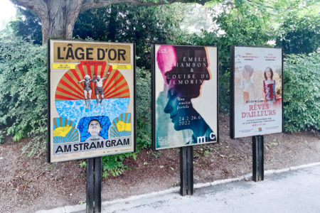 On a sidewalk in a park in Geneva, there are three billboards displaying various motifs from different theater productions.
