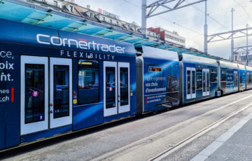 At a tram stop in Geneva, there is a tram with a blue full-cover motif advertising 'CornerTrader'