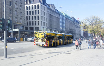 A bus in Hamburg is traveling with a full-cover motif featuring a yellow lion along a street.