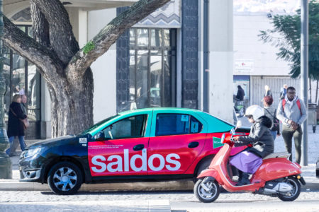 In Lisbon, a taxi is displaying advertising for 'Saldos,' with many people around.